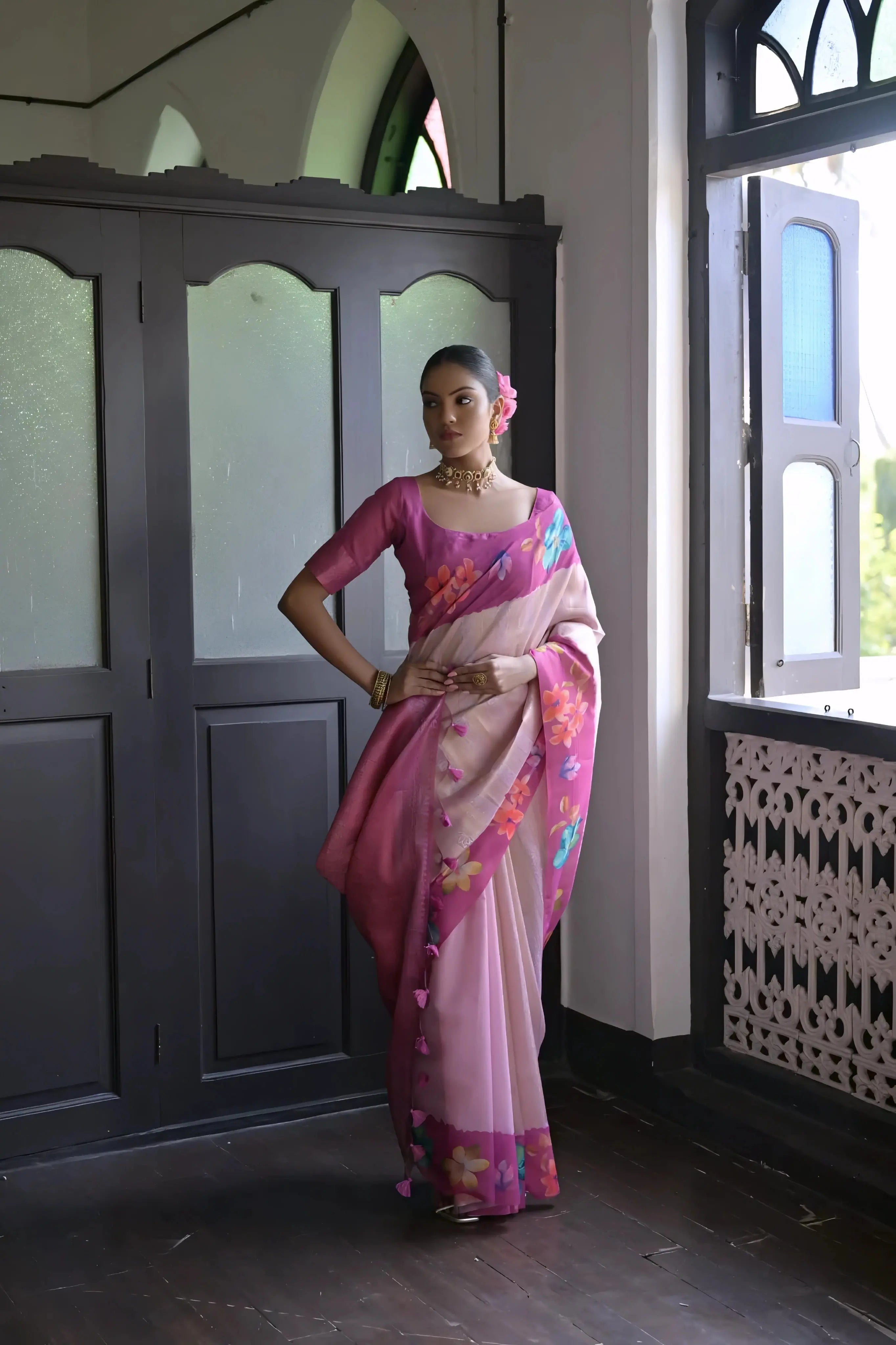 A woman models a graceful Soft Silk Saree. The saree body is light pink and features a wide, solid dark pink/fuchsia border. The shoulder and pallu area are decorated with large, vibrant printed floral motifs. She pairs it with a dark pink sheer-sleeve blouse and a gold choker necklace.